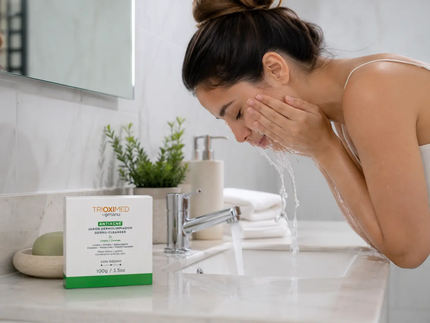 Rutina de limpieza facial con el Jab贸n Dermolimpiador Antiacn茅 Trioximed para piel grasa y con tendencia acneica. Mujer enjuagando su rostro en el lavabo con el Jab贸n Dermolimpiador Antiacn茅 Trioximed sobre el tocador del ba帽o durante su rutina de limpieza facial.